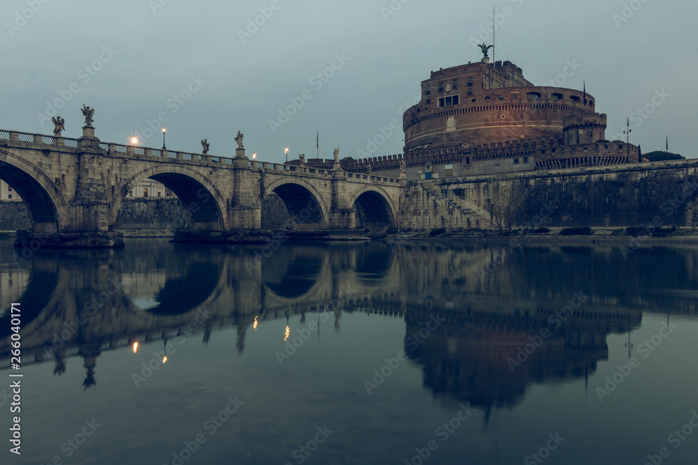 River Tiber with the Aurelius bridge over waters with Castel Sant Angelo at dusk. Reflections of the stone bridge in the water with illuminated buildings.