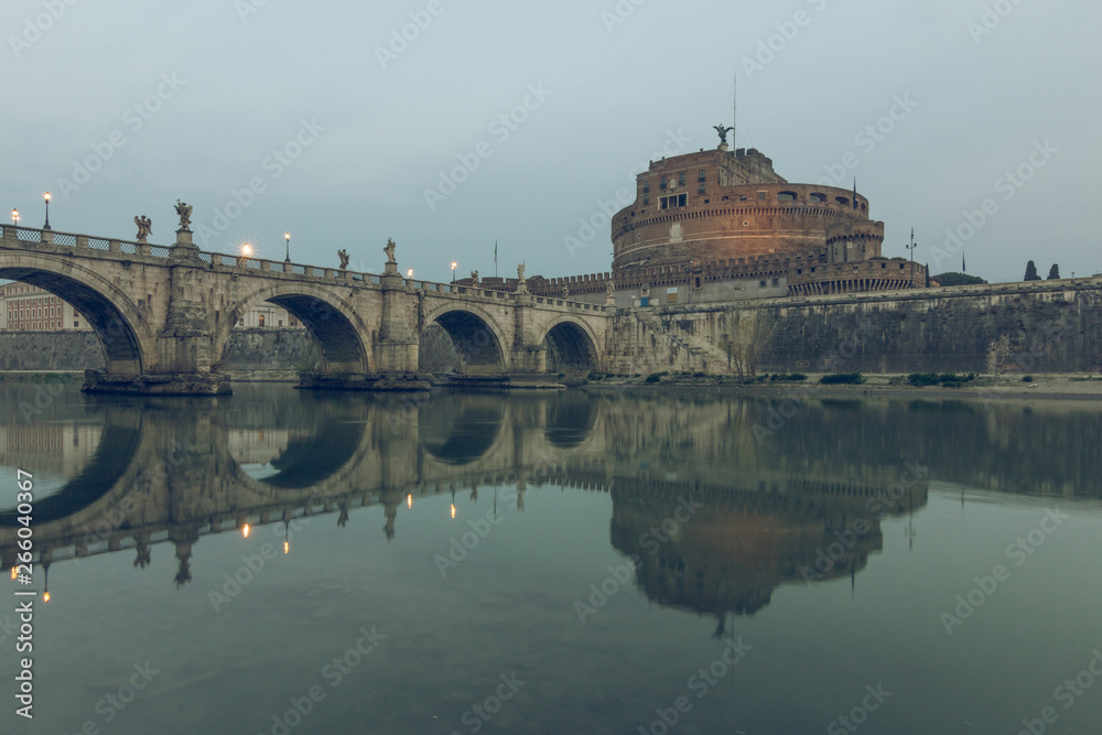 River Tiber in the historic center of Rome with the Aurelius bridge over waters with Castel Sant Angelo at dusk. Reflections of the stone bridge in the water with illuminated buildings