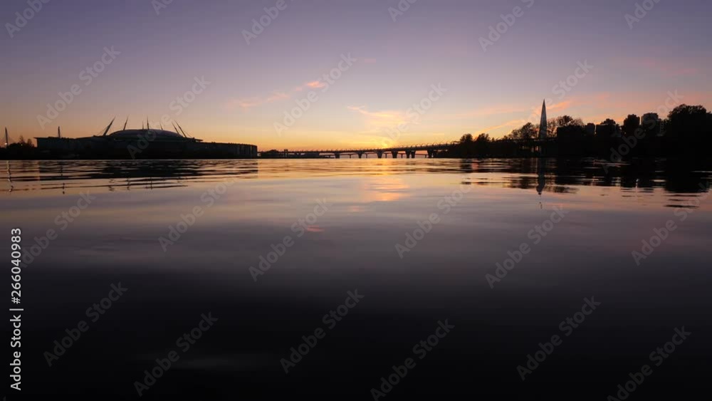 Small smooth waves on water surface, dark sunset sky reflection. Camera stay very low, close to quiet river water. Stadium building, road and tower, trees silhouetted on shores Small smooth waves on water surface, dark sunset sky reflection. Camera stay very low, close to quiet river water. Stadium building, road and tower, trees silhouetted on shores