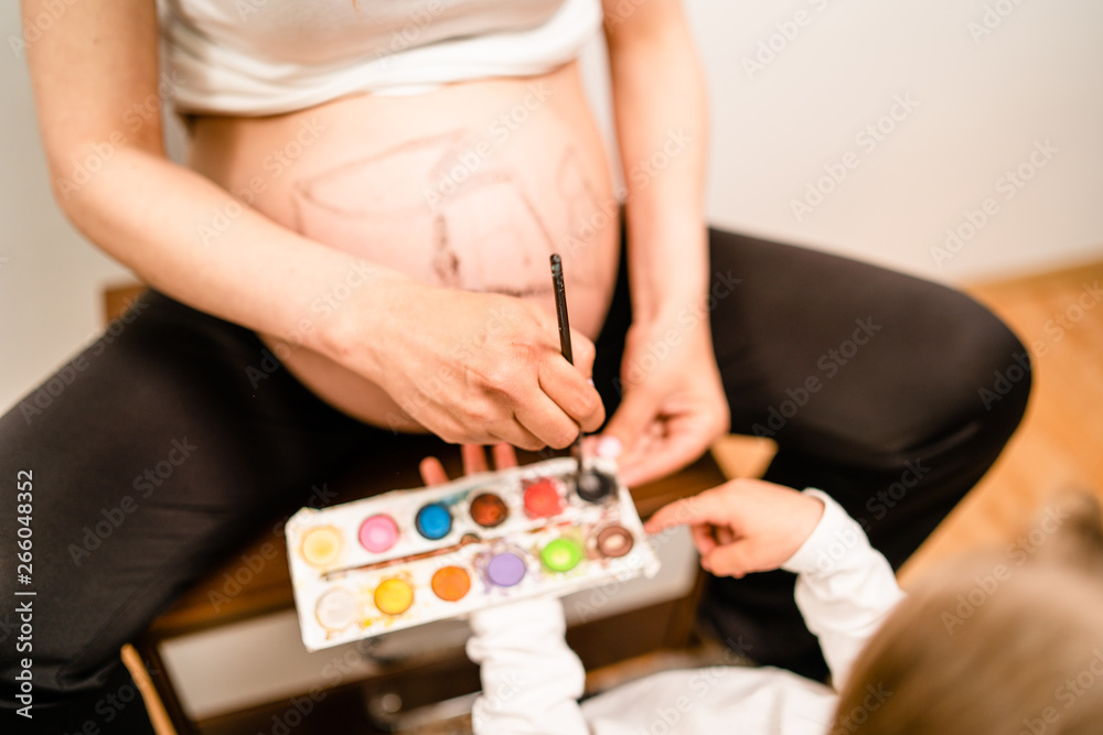 Little blonde girl painting little baby brother or sister on her mothers pregnant belly with water colors and paint brush Little blonde girl painting little baby brother or sister on her mothers pregnant belly with water colors and paint brush
