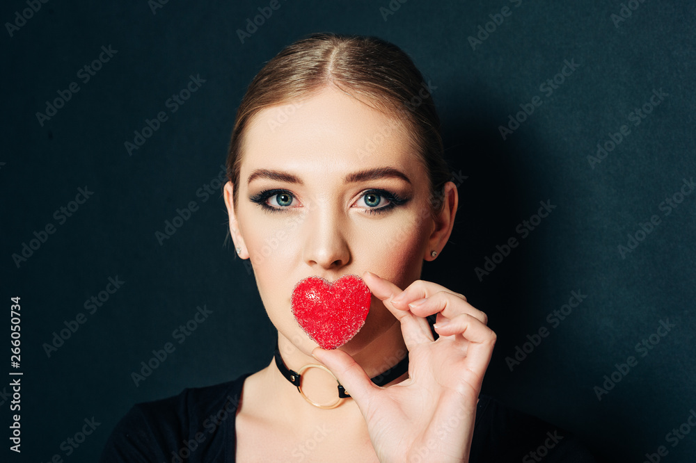 Close up studio portrait of beautiful young woman holding red heart next to mouth, black background Close up studio portrait of beautiful young woman holding red heart next to mouth, black background