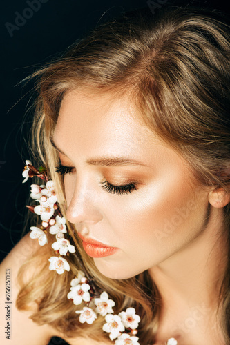 Close up portrait of beautiful young woman with blond hair and professional make up, posing on black background, holding spring blooming branch of peach flowers