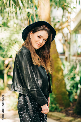 Outdoor portrait of beautiful young woman wearing black leather jacket and hat, green tropical leaves on background