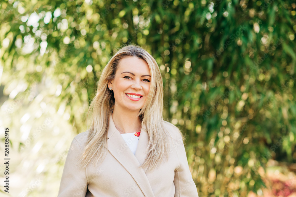 Outdoor portrait of beautiful blond woman wearing beige belted coat, posing in sunny garden Outdoor portrait of beautiful blond woman wearing beige belted coat, posing in sunny garden