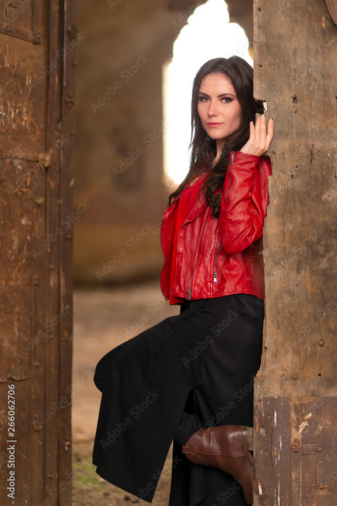 Fashion concept portrait of a gorgeous young woman sitting in a door of an old abandoned building and looking at the camera, in red leather jacket and black skirt outfit. Retouched, vibrant colors