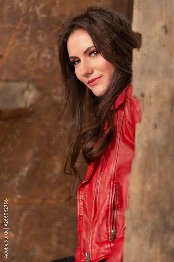 Fashion concept portrait of a gorgeous young woman in red leather jacket, sitting in a door of an old abandoned building, looking at the camera and smiling. Close-up shot, retouched for perfect skin Fashion concept portrait of a gorgeous young woman in red leather jacket, sitting in a door of an old abandoned building, looking at the camera and smiling. Close-up shot, retouched for perfect skin
