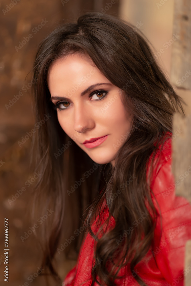 Naklejka premium Fashion portrait of a gorgeous young woman in red leather jacket, sitting in a door of an old abandoned building, looking at the camera. Close-up shot, retouched for perfect skin, vibrant colors