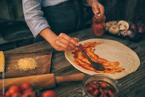 Fresh original Italian raw pizza and ingredients on wooden table, preparation in traditional style. Woman cooking homemade pizza on a wooden table in a home kitchen. Cooking, food and people concept.