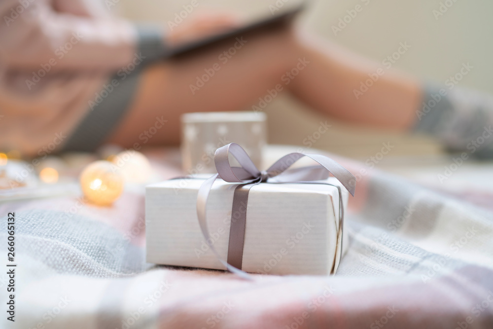 Close up white christmas gift box with pink ribbon, on the blurred cozy home background. Woman's hands typing on laptop keyboard. Warm pink sweater, winter, home comfort and relax. Toned image. Close up white christmas gift box with pink ribbon, on the blurred cozy home background. Woman's hands typing on laptop keyboard. Warm pink sweater, winter, home comfort and relax. Toned image.