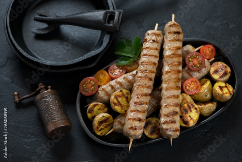 Cast-iron serving pan with barbecued chicken skewers, elevated view on a black stone background, horizontal shot