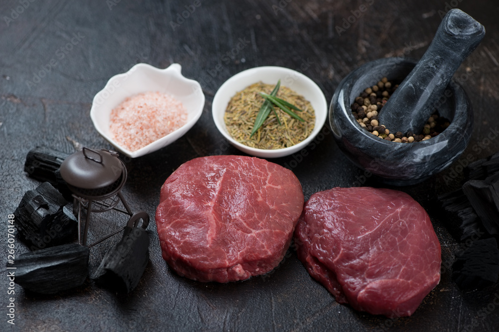 Raw fresh marbled beefsteaks with condiments, charcoal and a decorative grill on a dark brown stone background Raw fresh marbled beefsteaks with condiments, charcoal and a decorative grill on a dark brown stone background