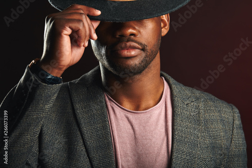 African cnfident business man, dressed in grey formal wear with black hat put down on eyes posing in studio on dark smoky background