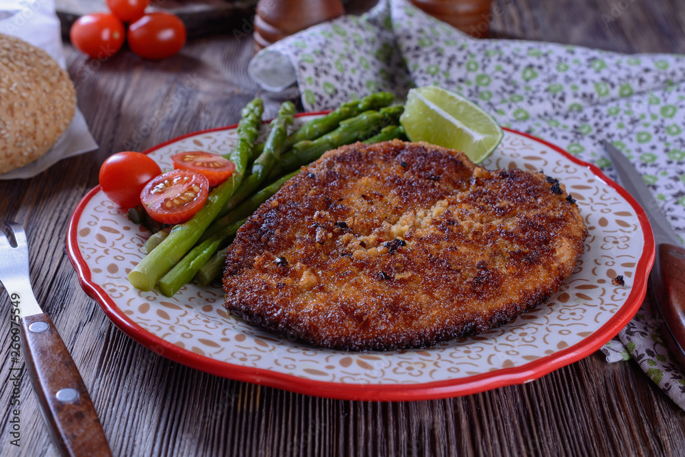Hot appetizing schnitzel with crispy crust on a plate next to asparagus, lime and cherry tomatoes on a wooden table are ready for eating Hot appetizing schnitzel with crispy crust on a plate next to asparagus, lime and cherry tomatoes on a wooden table are ready for eating