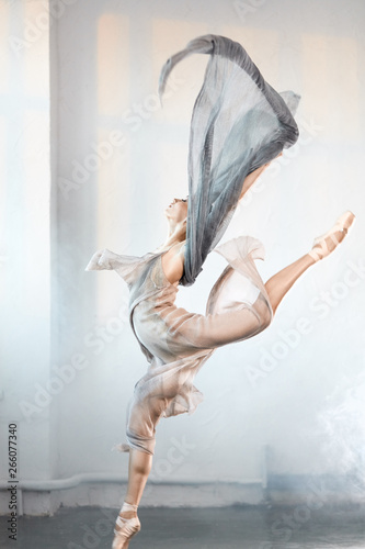 Fototapeta Naklejka Na Ścianę i Meble -  young ballet dancer wearing blue-grey transparent clothes and pointe has incredible grace, lightness and flexibility jumping on the stage with flying fabric and smoke effect