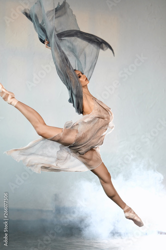 Fototapeta Naklejka Na Ścianę i Meble -  young ballet dancer wearing blue-grey transparent clothes and pointe has incredible grace, lightness and flexibility jumping on the stage with flying fabric and smoke effect