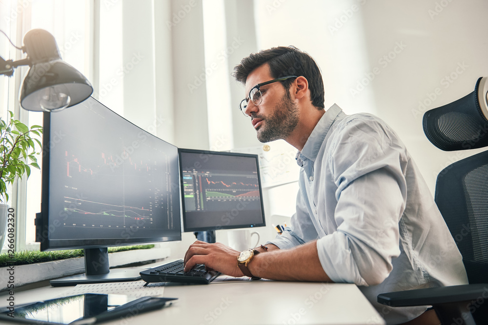 Confident businessman. Young bearded trader in formal wear is analyzing trading charts on computer screens while sitting in his modern office - obrazy, fototapety, plakaty 