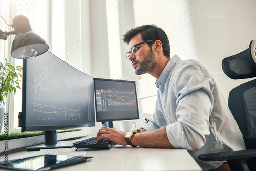 Confident businessman. Young bearded trader in formal wear is analyzing trading charts on computer screens while sitting in his modern office