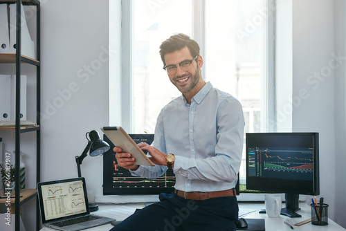 Business success. Cheerful young trader in formal wear is holding digital tablet and looking at camera with smile while standing in front of computer screens with trading charts in the office. photo