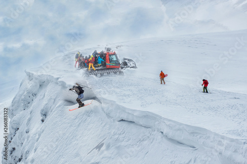 A group of freeriders arrived at the top of the mountain on a snowmobile. One skier jumps off a cliff photo