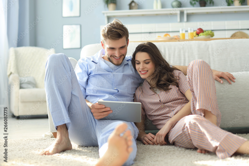 Obraz premium Young couple in pajamas watching media content online in a tablet sitting on the floor in the living room.