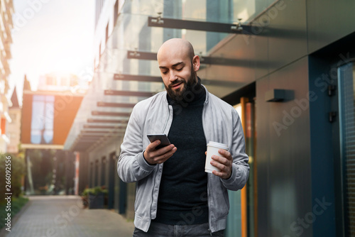 Portrait of young smiling man using smartphone on city street. Man sends text message, drinks coffee. Lifestyle. Social networks.