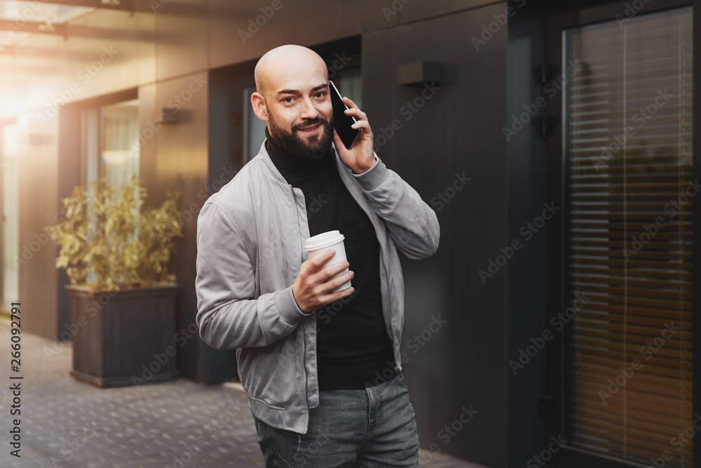Portrait of smiling man talking on cell phone and drinking coffee on city street, bearded hipster guy calling friends. Lifestyle. Portrait of smiling man talking on cell phone and drinking coffee on city street, bearded hipster guy calling friends. Lifestyle.