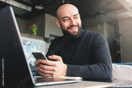 Bald bearded man in black turtleneck working on laptop,holding smartphone while sitting in cafe at table.Freelancer works