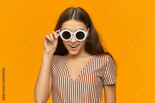 Style, fashion and positive emotions concept. Portrait of happy joyful young lady wearing stylish sunglasses in white frame and striped top smiling broadly at camera, going to have fun at party