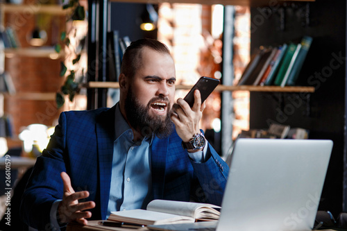 Serious bearded businessman conducts telephone conversations on a mobile phone at the computer. Unhappy man scolds on the phone. Emitations of rage in the office at the manager