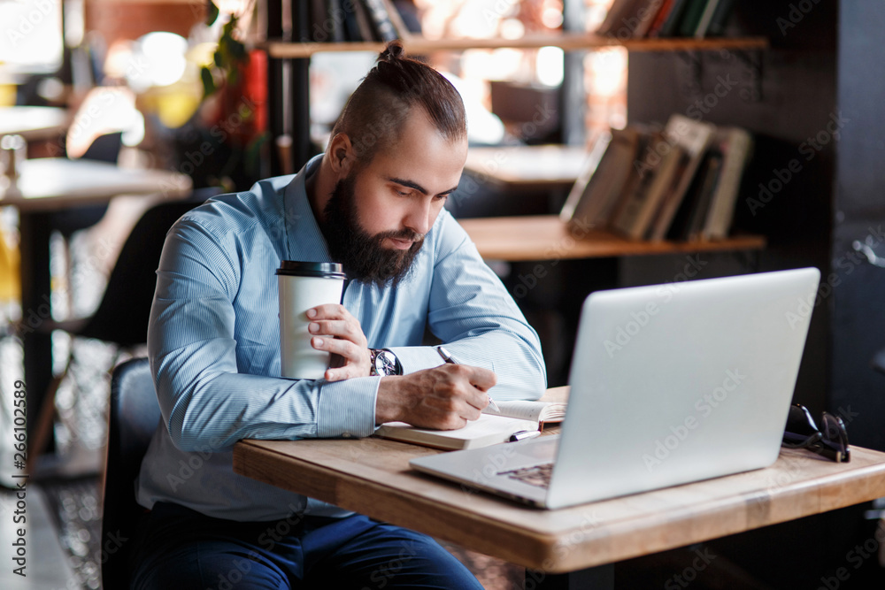 Young serious bearded businessman working on computer at table,drinking coffee.Man analyzes information, data, develops business plan. Freelancer, entrepreneur.Online marketing, education, e-learning
