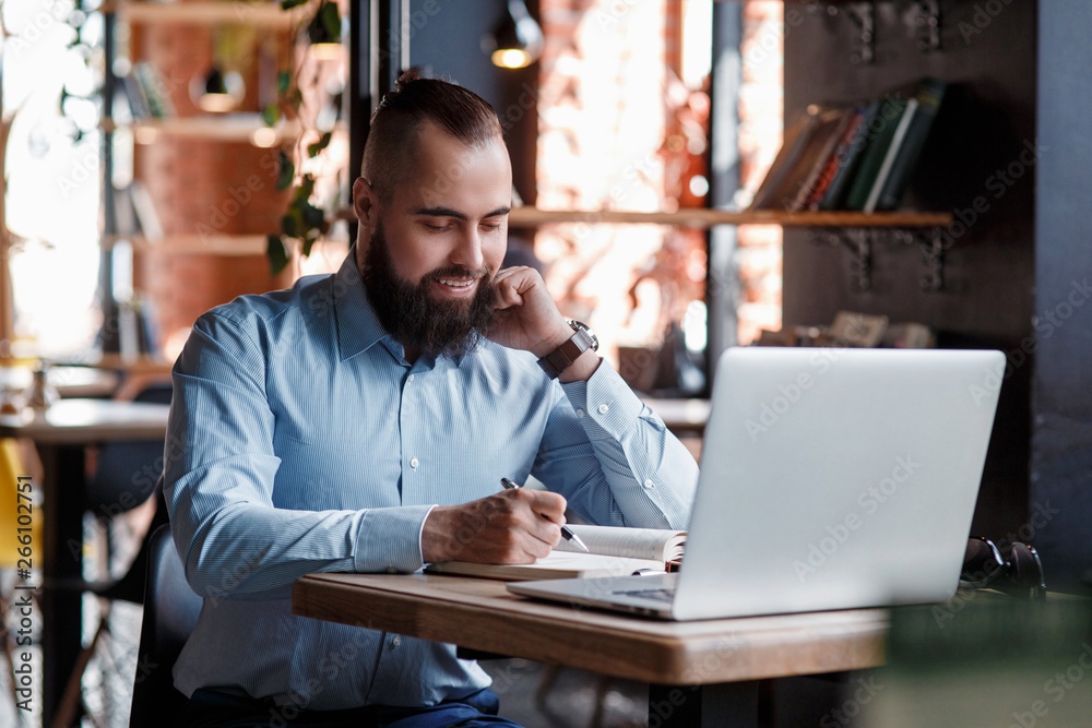 Young serious bearded businessman working on computer at table,drinking coffee.Man analyzes information, data, develops business plan. Freelancer, entrepreneur.Online marketing, education, e-learning