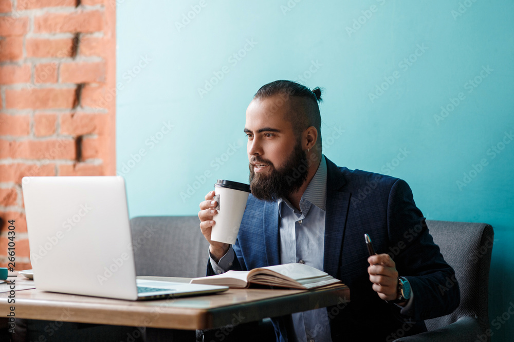 Young serious bearded businessman working on computer at table,drinking coffee.Man analyzes information, data, develops business plan. Freelancer, entrepreneur.Online marketing, education, e-learning Young serious bearded businessman working on computer at table,drinking coffee.Man analyzes information, data, develops business plan. Freelancer, entrepreneur.Online marketing, education, e-learning