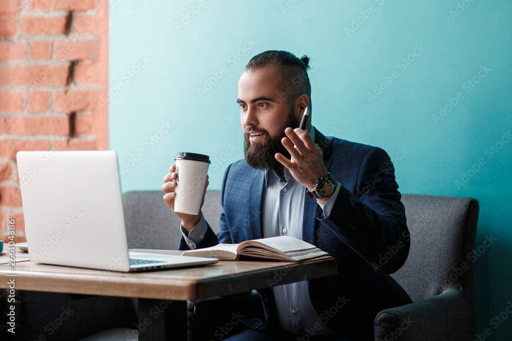 Young serious bearded businessman working on computer at table,drinking coffee.Man analyzes information, data, develops business plan. Freelancer, entrepreneur.Online marketing, education, e-learning Young serious bearded businessman working on computer at table,drinking coffee.Man analyzes information, data, develops business plan. Freelancer, entrepreneur.Online marketing, education, e-learning