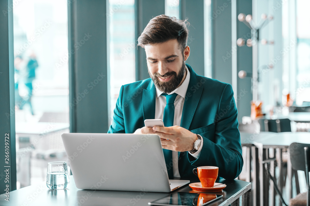 Smiling attractive ambitious businessman in turquoise suit using smart phone for reading or writing message while sitting in cafe. On desk glass of water, laptop, coffee and tablet. Smiling attractive ambitious businessman in turquoise suit using smart phone for reading or writing message while sitting in cafe. On desk glass of water, laptop, coffee and tablet.