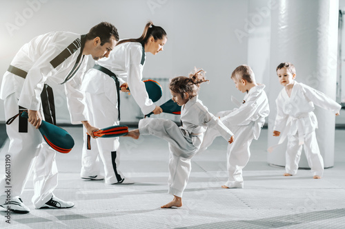 Small group of kids in doboks practicing with their trainers taekwondo moves while kicking in kick target.