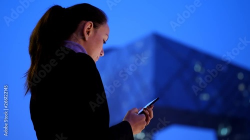 Woman texting in smartphone standing against night modern cityscape