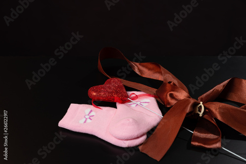 Baby pink socks with a bow and with a red heart are lying on a black table, on a black background.