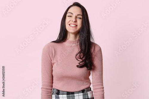 Photo of happy young woman with beautiful dark long hair laughing isolated over pink background, having fun with her friends, hearing jokes, expresses happyness, being glad to be photographed.