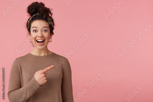 Portrait of happy amazed young beautiful lady with curly dark hair, heard the cool news, broadly smiling, looking at the camera, pointing with finger to copy space isolated over pink background. photo