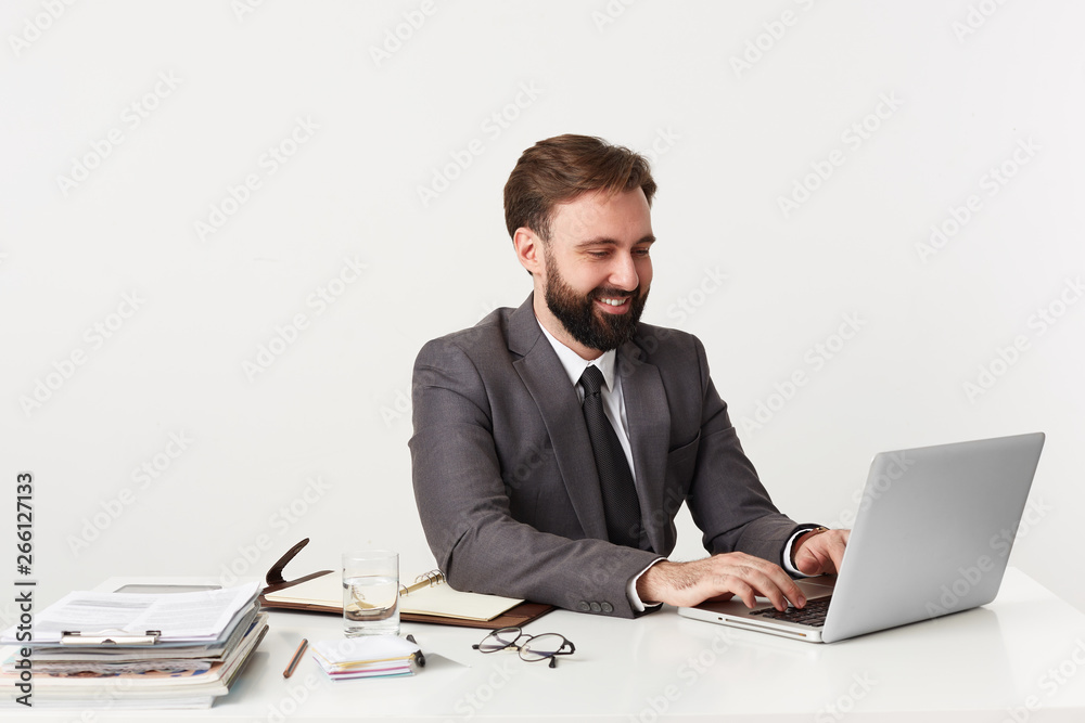 Glad smiling attractive bearded businessman, top manager sitting at desktop in office, working for his laptop , dressed in an expensive suit with a tie, enjoy his work. Glad smiling attractive bearded businessman, top manager sitting at desktop in office, working for his laptop , dressed in an expensive suit with a tie, enjoy his work.