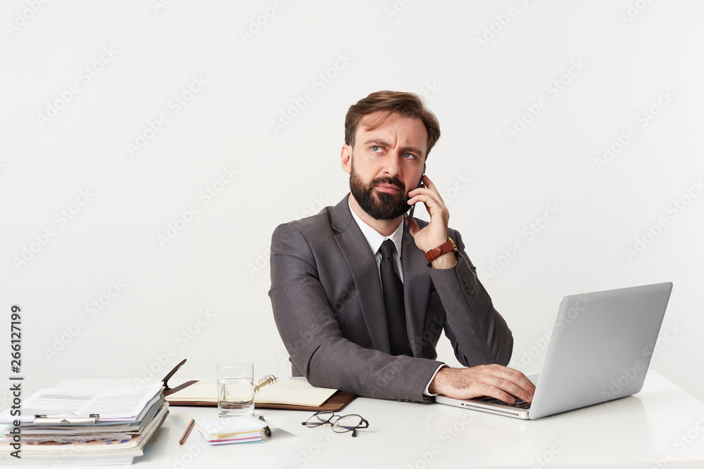 Young attractive businessman looking up thoughtfully and talking on the phone, trying to come up with a solution. Sitting at desktop in office, working for his laptop, dressed in a suit with tie. Young attractive businessman looking up thoughtfully and talking on the phone, trying to come up with a solution. Sitting at desktop in office, working for his laptop, dressed in a suit with tie.