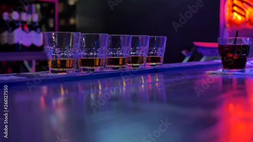 on a bar counter in a dark room there are five glasses with alcohol and one with a cocktail and a straw. The girl the bartender reached out for a drink