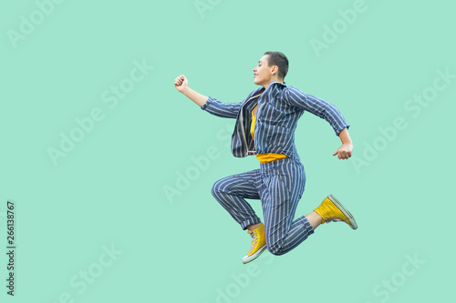 Profile side view portrait of happy handsome beautiful short hair young stylish woman in casual striped suit jumping in super mario pose. indoor studio shot isolated on light green background.