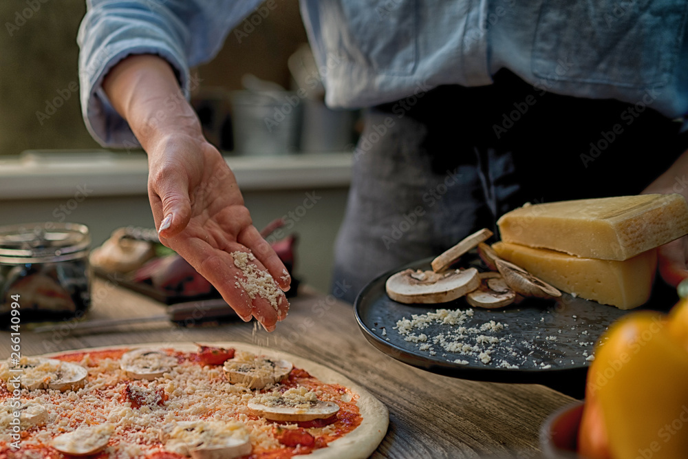 A housewife put chees on base in a rural italian kitchen. Make pizza for dinner, hands add ingriduent. Italian food making pizza with ingredients, tomatoes, chees and mushrooms on wooden tabletop.