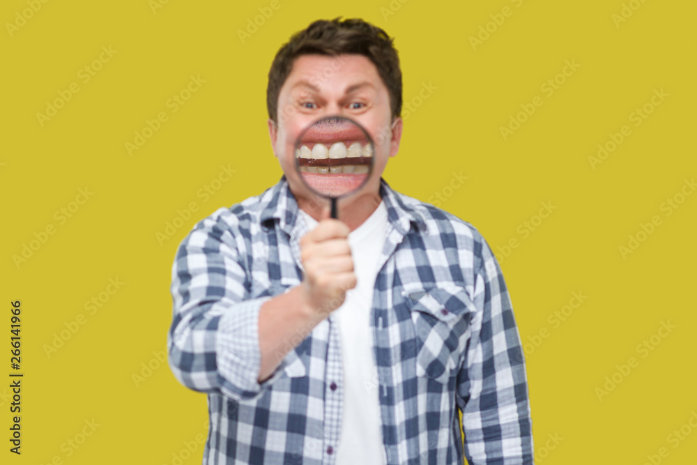 smile zoom or tooth care concept. Portrait of middle aged man in casual checkered shirt standing, holding magnifying glass on his teeth, looking at camera. studio shot, isolated on yellow background. smile zoom or tooth care concept. Portrait of middle aged man in casual checkered shirt standing, holding magnifying glass on his teeth, looking at camera. studio shot, isolated on yellow background.