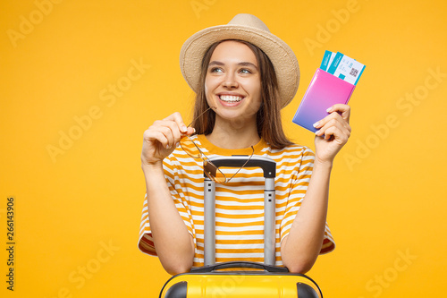 Close-up studio portrait of dreaming young female tourist holding flight tickets and suitcase, isolated on yellow background