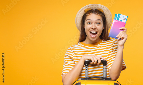 Horizontal banner of happy surprised young female tourist  holding suitcase passport with flight tickets, isolated on yellow background with copy space photo