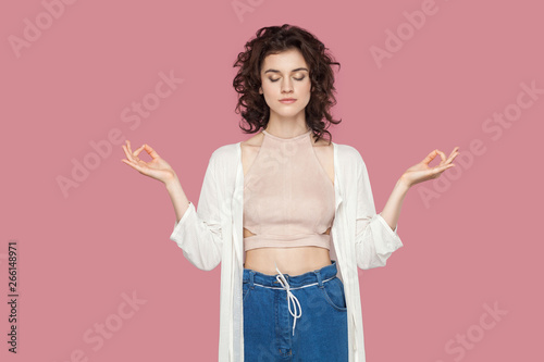 Portrait of calm relaxed beautiful brunette young woman with curly hairstyle in casual style standing in yoga pose raised arms, closed eyes, meditating. indoor studio shot isolated on pink background.