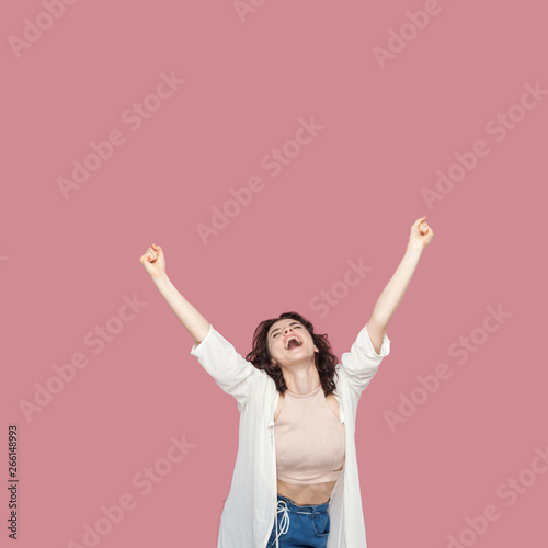Portrait of happy satisfied beautiful brunette young woman with curly hairstyle in casual style standing, screaming and celebrating her victory, raised arms. studio shot isolated on pink background.