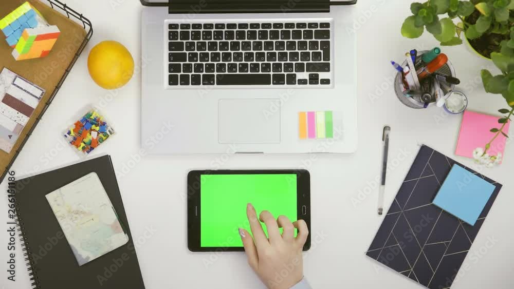 Top view on the office desk and white tablet computer with green screen and female hands taping on it.